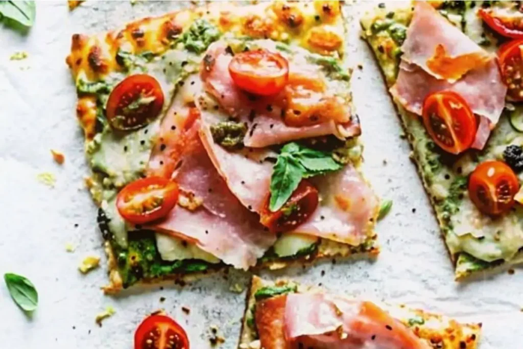 Low carb pesto chicken crust pizza slices topped with cherry tomatoes, ham, mozzarella, and fresh basil