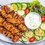 Grilled Shish Tawook chicken skewers served with cucumber, tomato salad, lime slices, and creamy yogurt garlic sauce on a white platter